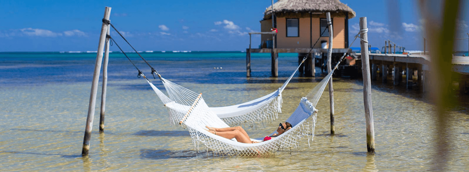 A woman relaxes in a hammock over clear water with a thatched-roof building and blue sky in the background.