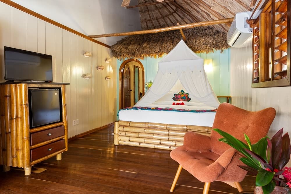 A bedroom with a king bed covered by a mosquito net, a thatched roof section, an coral armchair, a TV, and wooden floors.