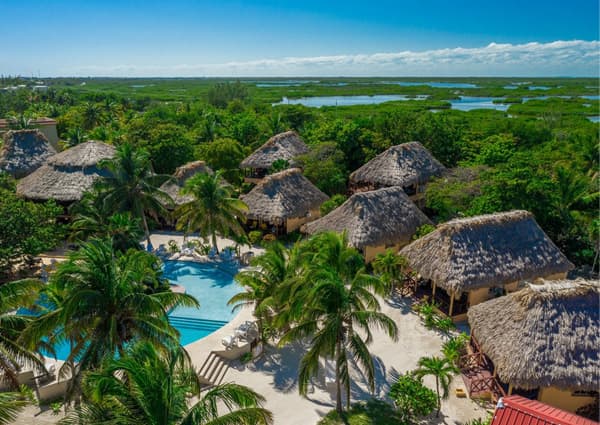 Aerial view of a tropical resort featuring thatched-roof huts, a swimming pool, and lush greenery near a waterway.