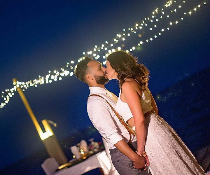 A couple shares a romantic kiss under twinkling lights by the water at dusk.