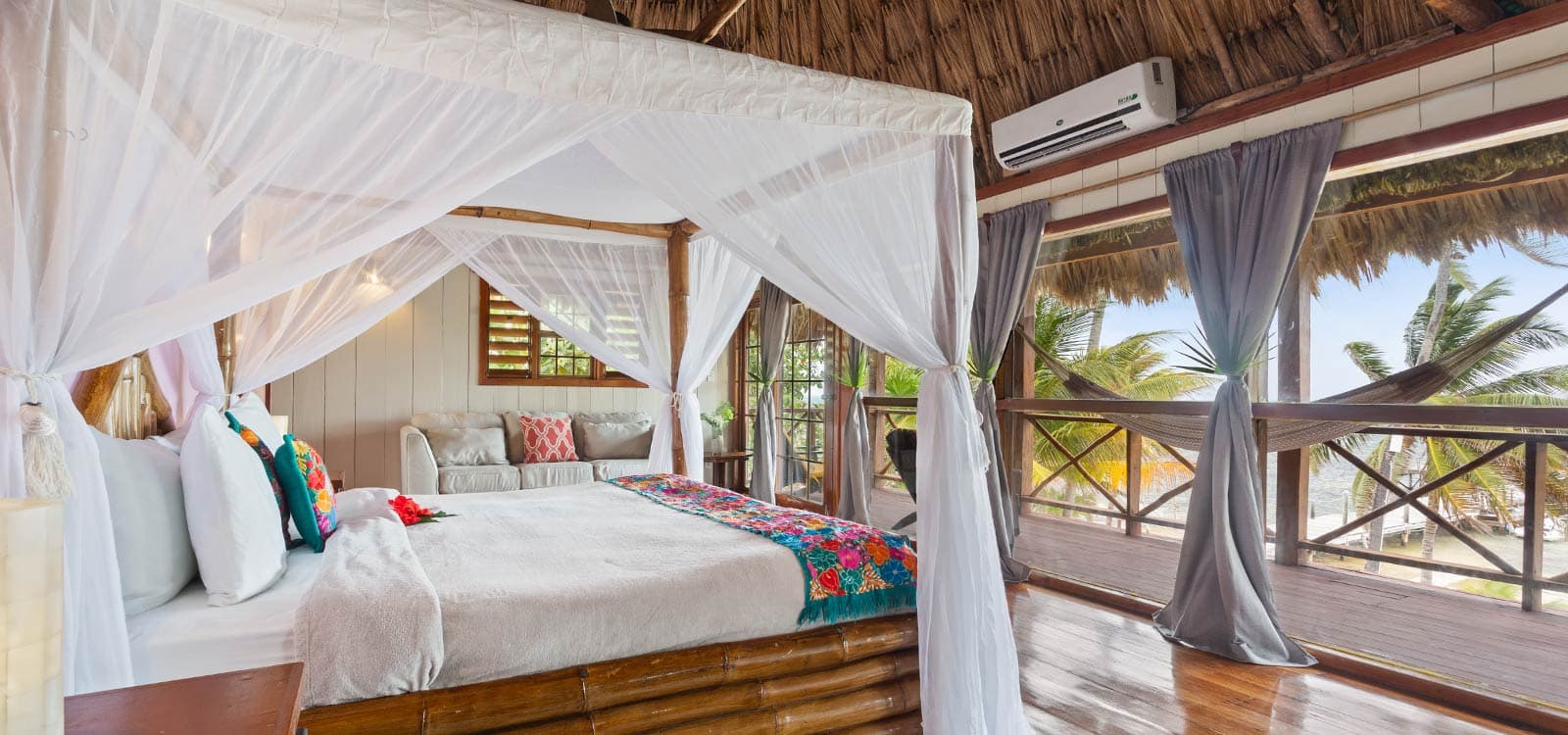 A cozy bedroom with a canopy bed, white curtains, and a view of palm trees and the ocean.