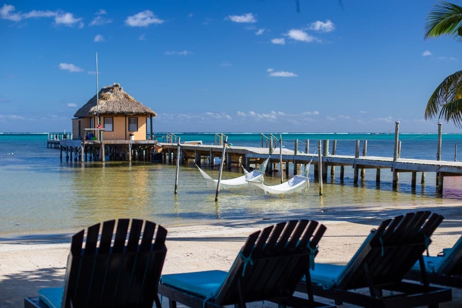 A thatched-roof hut overlooks a calm turquoise sea, with two hammocks hanging between posts and beach chairs in the foreground.