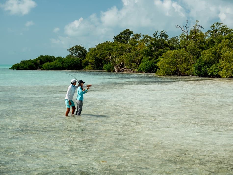 Two anglers wade through shallow, clear waters surrounded by lush mangroves.