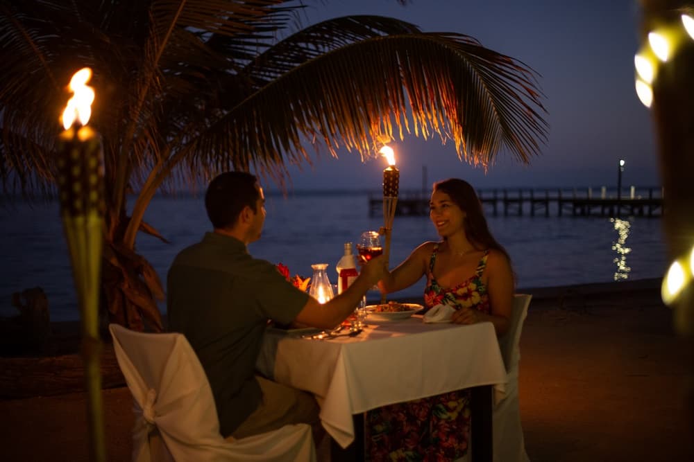 A couple toasts over a romantic dinner at sunset by the water, illuminated by tiki torches.