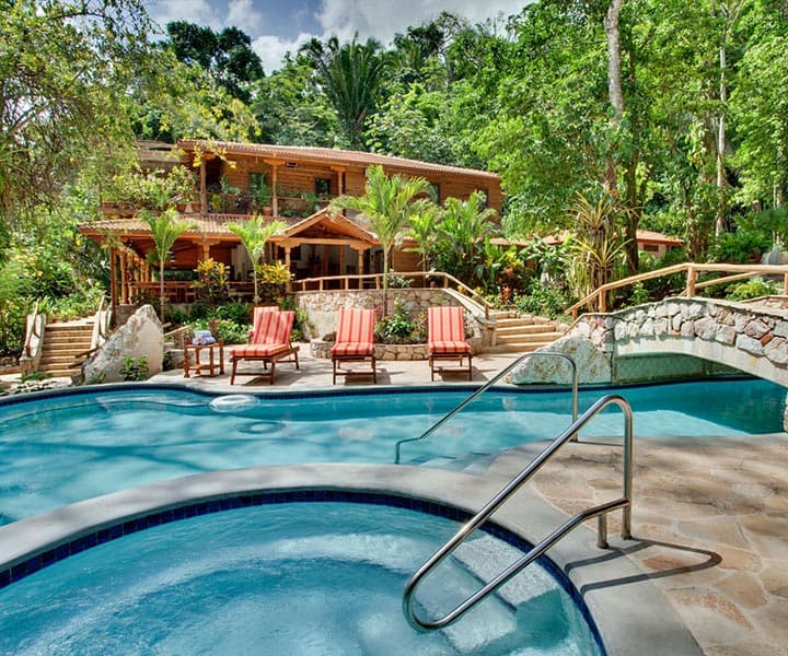 A serene pool area with red lounge chairs, surrounded by lush greenery and a wooden house in the background.