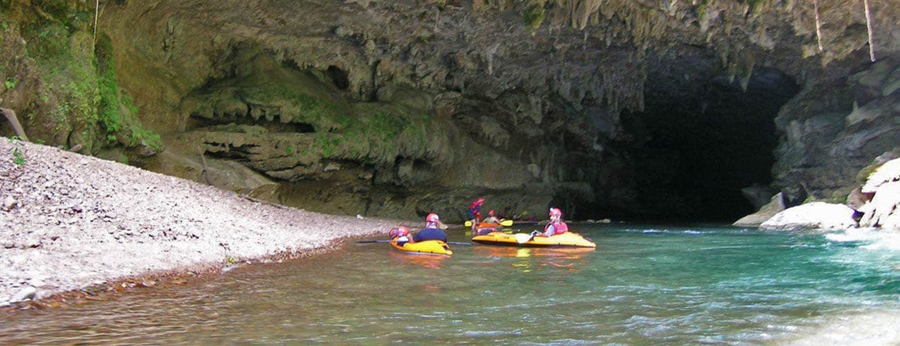 Kayakers navigate a serene river near a rocky cave entrance.