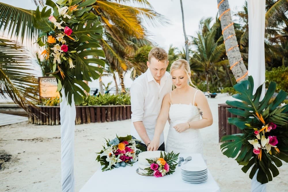 A newly married couple cuts their wedding cake on a beach, surrounded by tropical decorations.