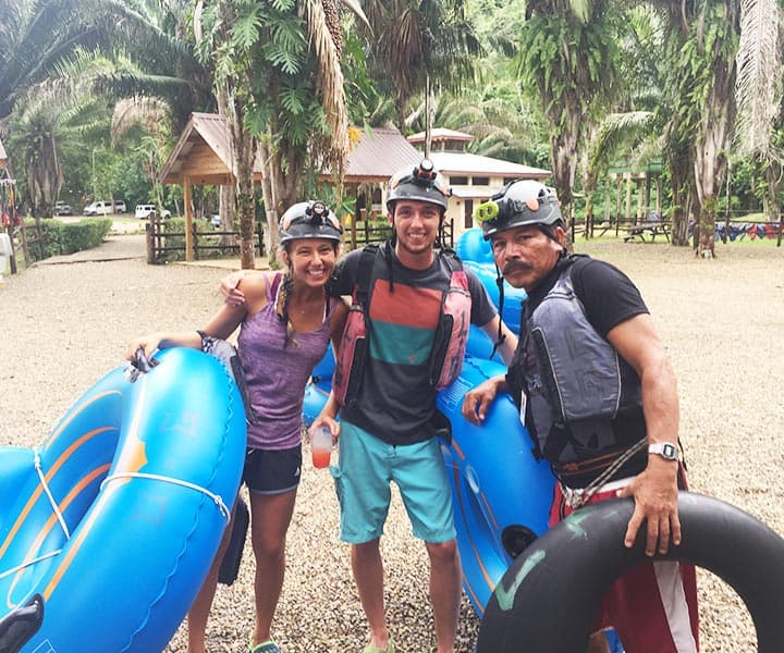 Three people with helmets and life jackets pose with inflatable rafts in a tropical outdoor setting.