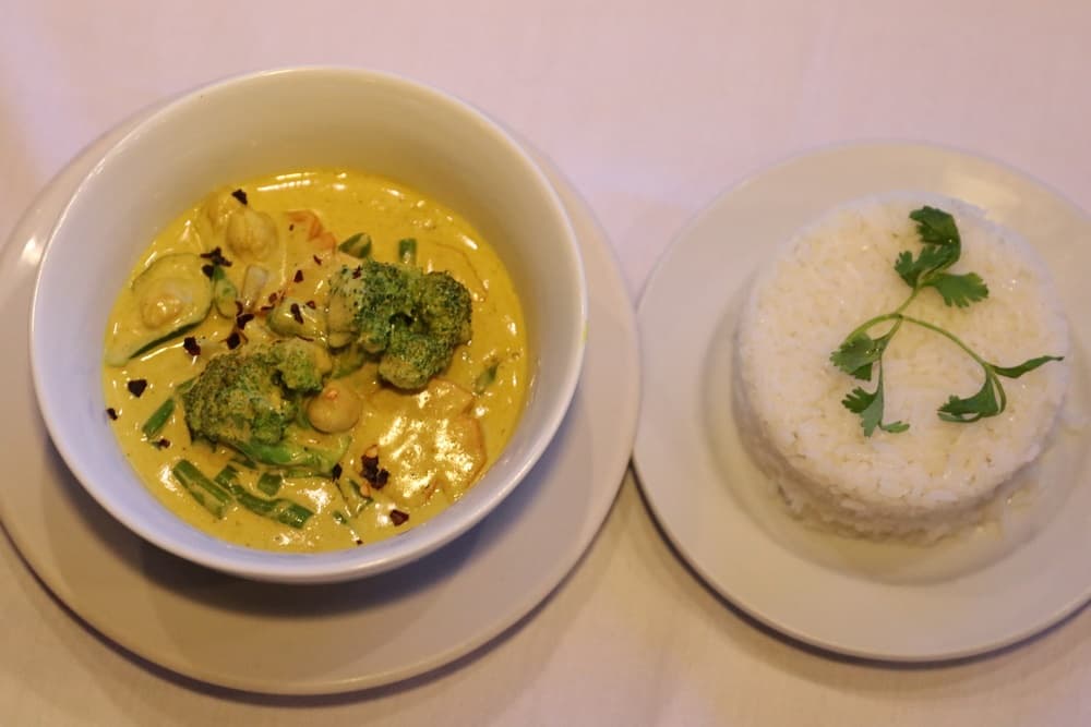 A bowl of creamy curry with broccoli and veggies alongside a serving of fluffy white rice garnished with a sprig of cilantro.