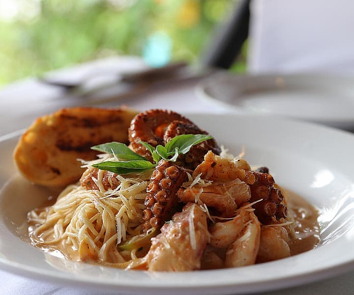A plate of seafood pasta topped with octopus, shrimp, and fresh basil, served with a side of toasted bread.