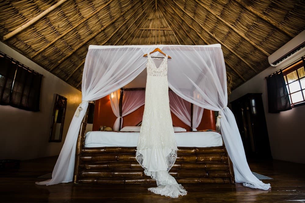 A wedding dress hangs gracefully from a wooden hanger in a beautifully decorated room with a thatched roof.