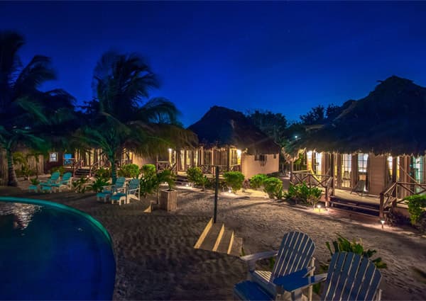 A serene beach resort with thatched-roof cabins illuminated at night, surrounded by palm trees and a pool.