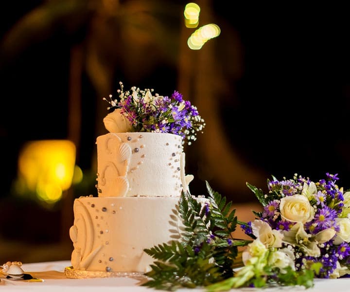 A three-tiered wedding cake adorned with flowers and greenery, set against a softly lit background.