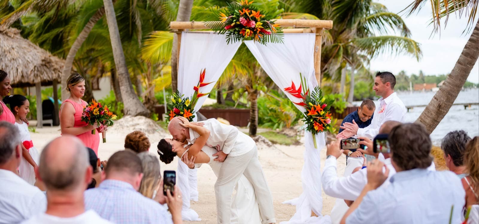 A couple shares a joyous kiss under a tropical wedding arch adorned with vibrant flowers. Guests in casual attire capture the moment with phones, surrounded by palm trees and a beach backdrop.