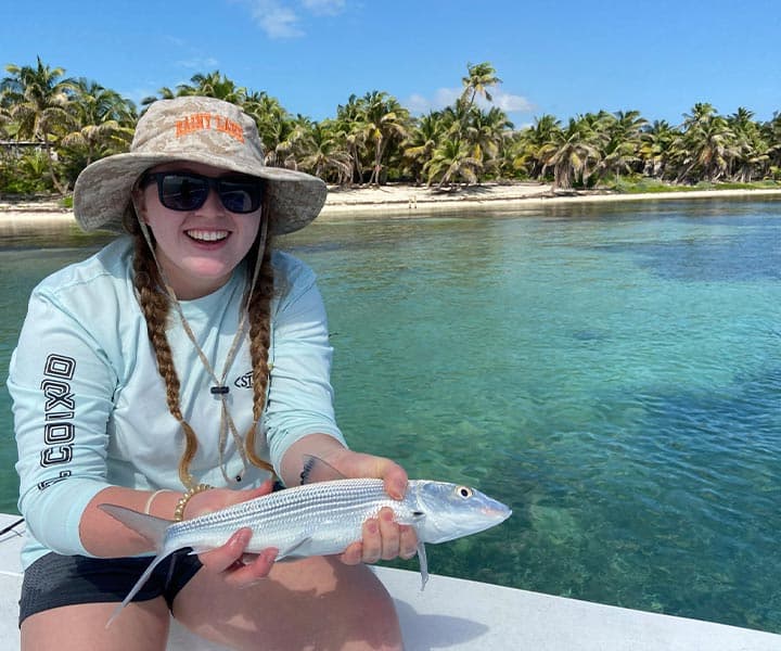 A smiling woman in a hat holds a fish while sitting on a boat with a tropical background.