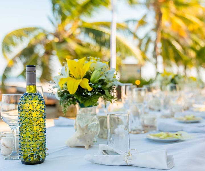 A beautifully set outdoor dining table featuring a decorated wine bottle, a floral centerpiece, and palm trees in the background.