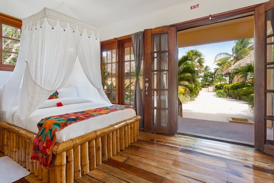 A bright, tropical bedroom features a bamboo bed with a white canopy, opening to a lush outdoor view.