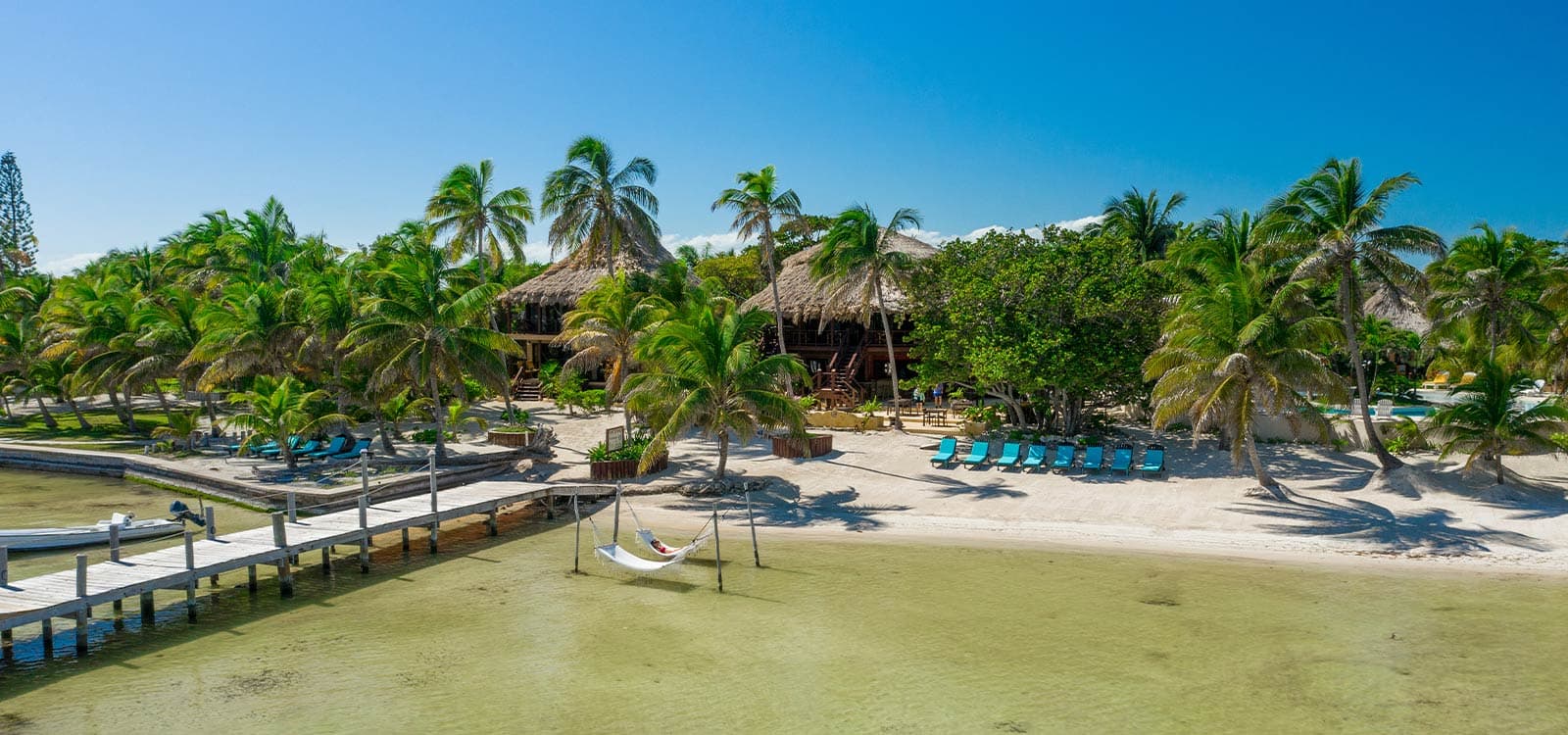 Tropical beachfront scene featuring palm trees, hammocks, and thatched-roof cabins under a clear blue sky.