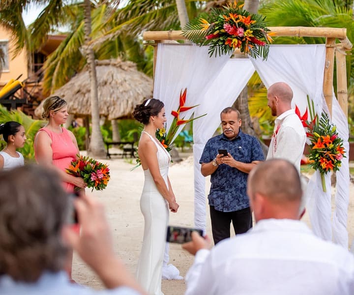 A couple exchanges vows at a tropical beach wedding ceremony with attendees holding flowers.