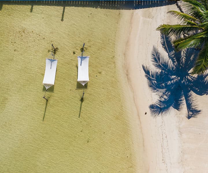 Aerial view of two beach loungers on a sandy shore with a palm tree shadow.