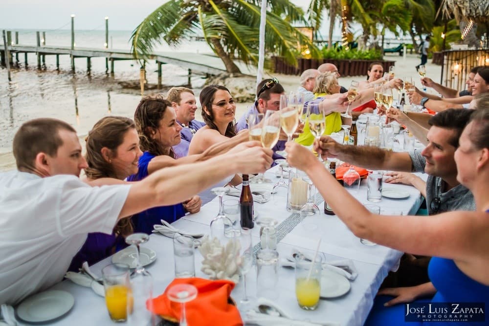A group of people toasting with drinks at a festive outdoor dining table by the beach.