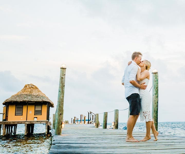 A couple kisses on a wooden pier over the water, with a thatched-roof hut in the background.