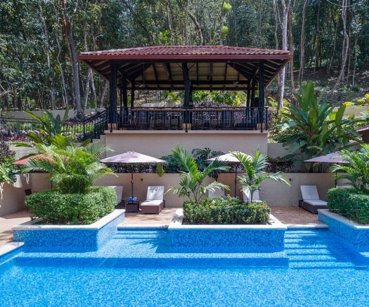 A serene pool area with lush greenery and a covered pavilion in the background.