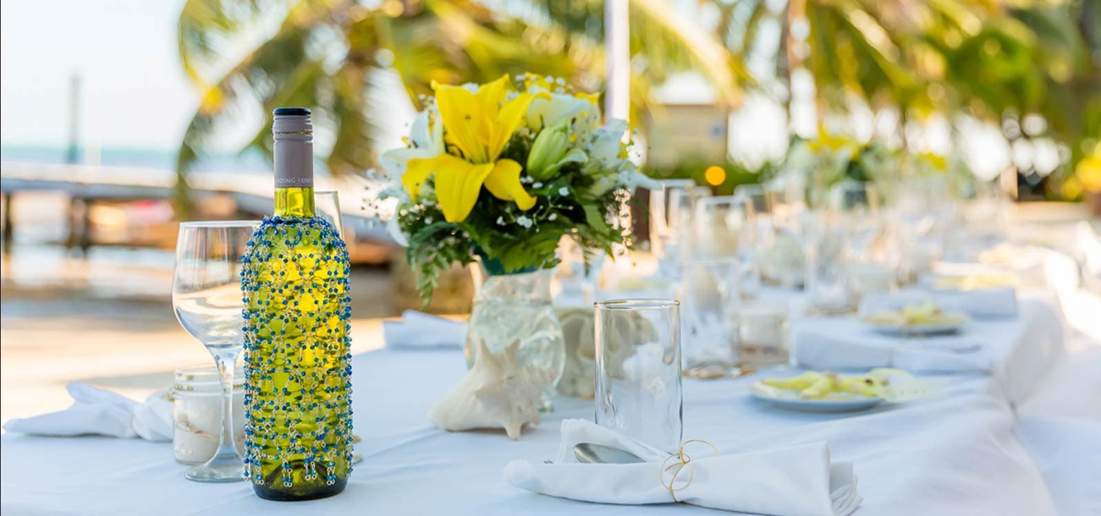 A decorated wine bottle and glass sit on a white-clothed table adorned with yellow lilies and seashells, framed by palm trees, evoking a tropical, elegant setting.