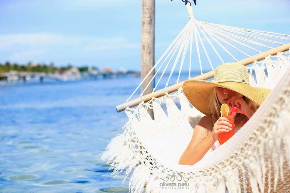 A woman relaxes in a hammock by the water, sipping a colorful drink under a wide-brimmed hat.