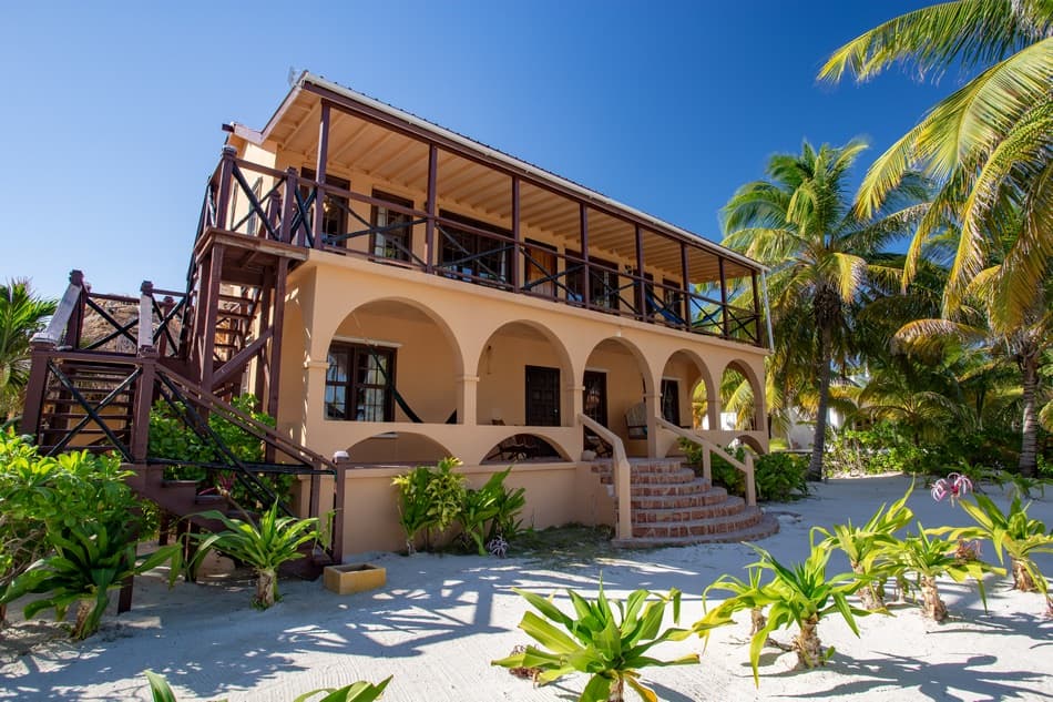 A two-story beach house with arched doorways and surrounded by lush greenery and palm trees.