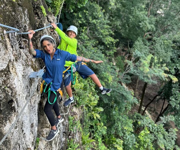 Two climbers navigate a rock face while harnessed and secured, surrounded by lush greenery.