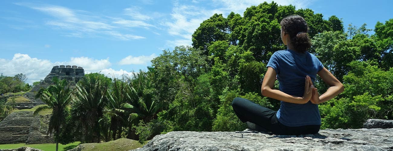 A person sits cross-legged on a rock, meditating with hands behind their back, in front of lush greenery and Mayan ruins. The scene feels tranquil and serene.
