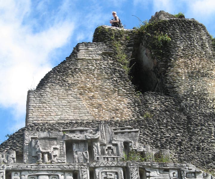 A person sits atop a stone pyramid, surrounded by lush greenery and a blue sky.