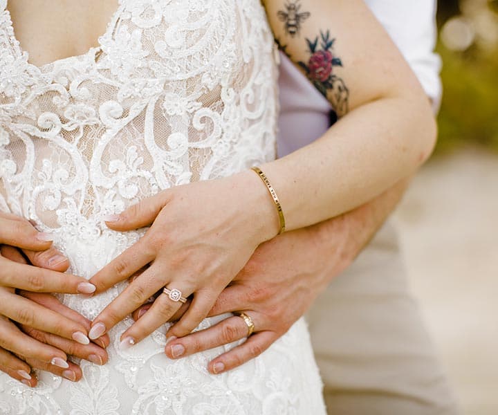 A close-up of intertwined hands displaying rings and a tattoo on a bride's arm, set against a wedding dress.