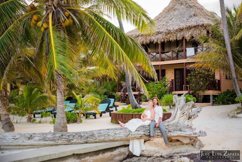 A couple poses joyfully on a driftwood log in front of a tropical beach house surrounded by palm trees.