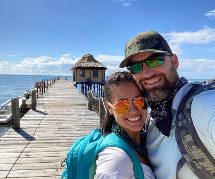 A couple takes a selfie on a sunny day by a wooden pier leading to a thatched-roof hut over the water.
