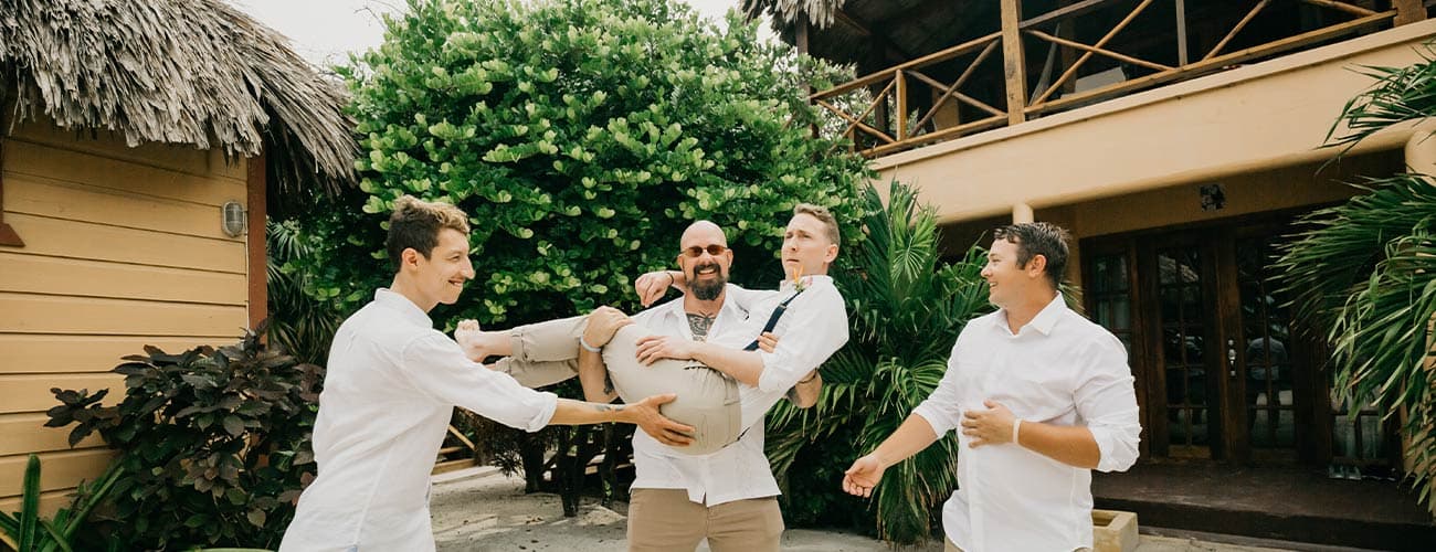 Four men in white shirts playfully carry one another outside a tropical building.