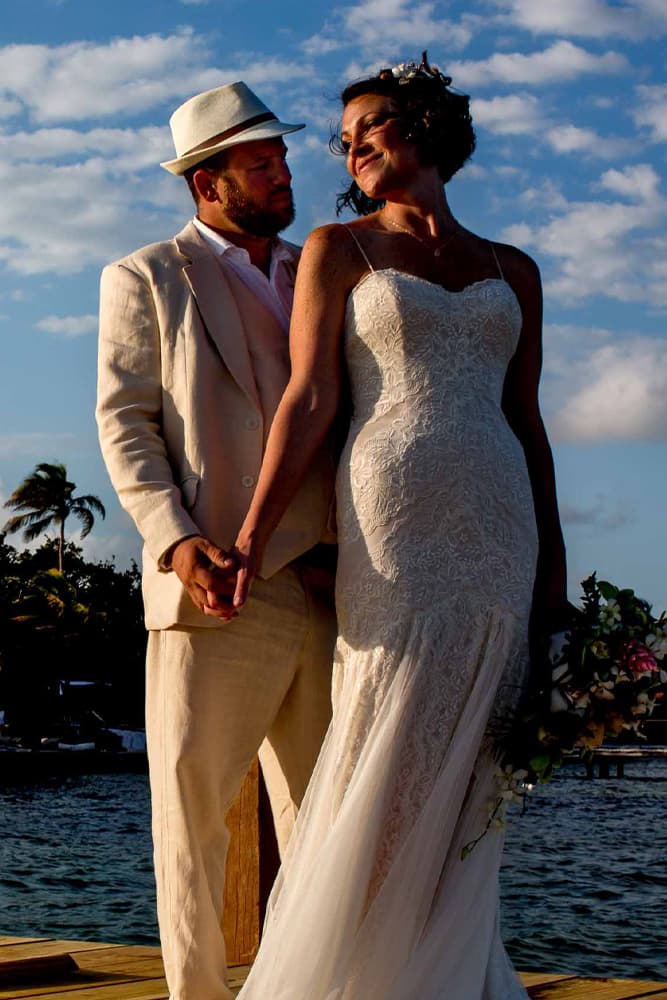 Bride and groom stand on a dock at sunset, holding hands and gazing at each other. The bride wears a lace gown; the groom, a light suit and hat. Romantic ambiance.