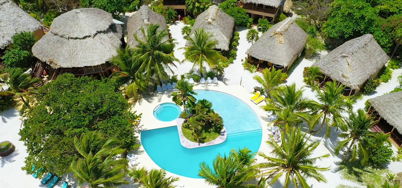 Aerial view of a tropical resort with thatched-roof bungalows surrounding a large, blue pool. Palm trees and lush greenery create a serene, paradise-like ambiance.