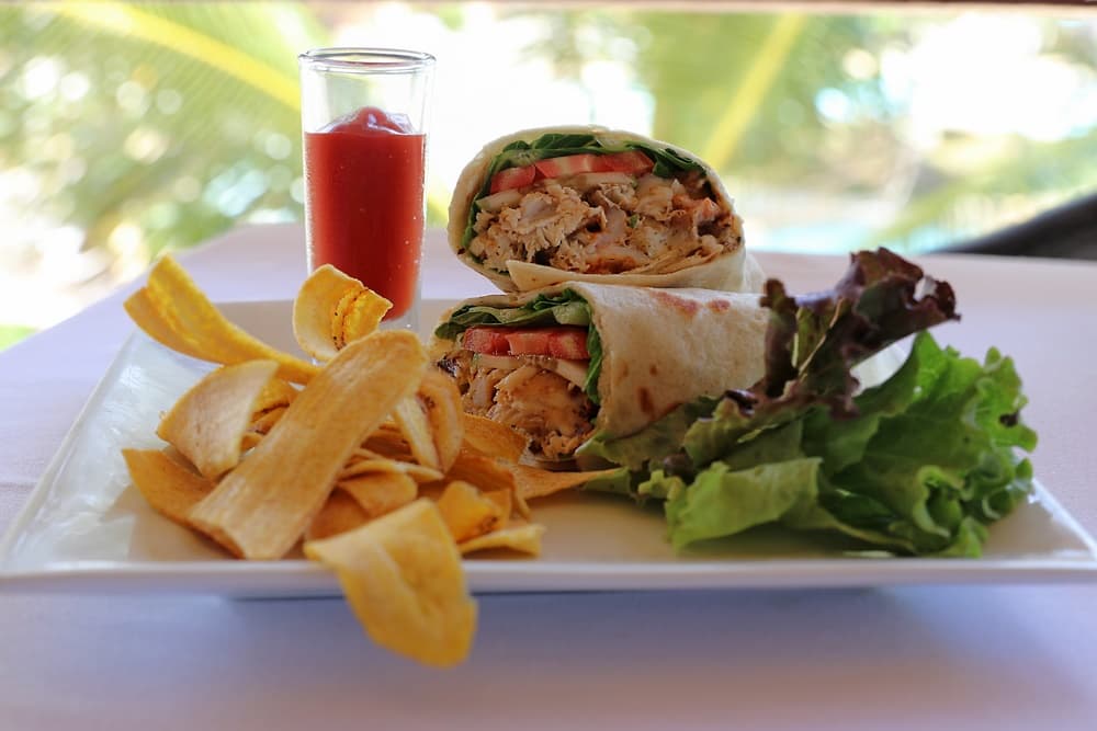 A plate featuring two chicken wraps with lettuce and tomato, served alongside plantain chips and a small glass of sauce.