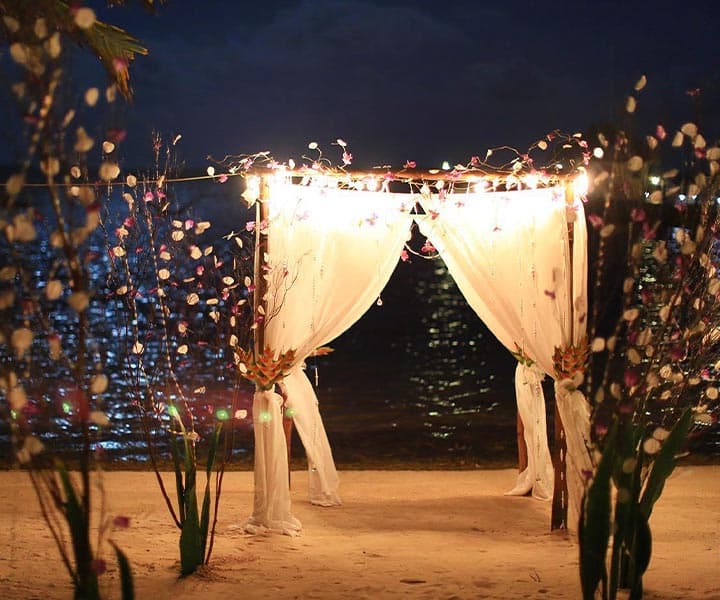 A beautifully lit ceremonial arch draped in white fabric and surrounded by flowers at the water's edge during the evening.
