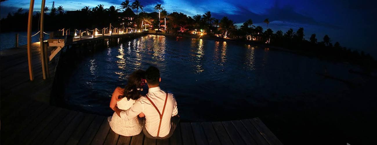 A couple sits together on a dock, overlooking a tranquil waterway illuminated by evening lights.