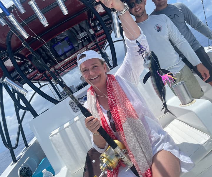A woman in a white outfit and cap smiles while holding a fishing rod with a caught fish on a boat.