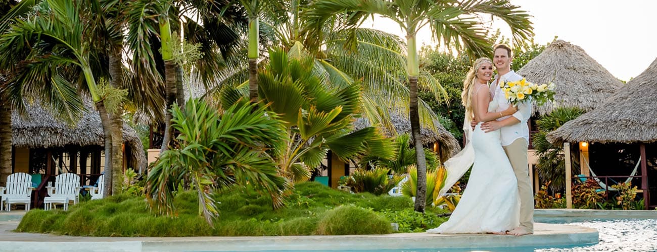 A bride and groom embrace by a pool surrounded by tropical plants and thatched-roof huts.