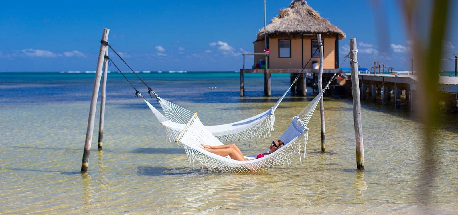 A person relaxes in a hammock over shallow water with a tropical backdrop.
