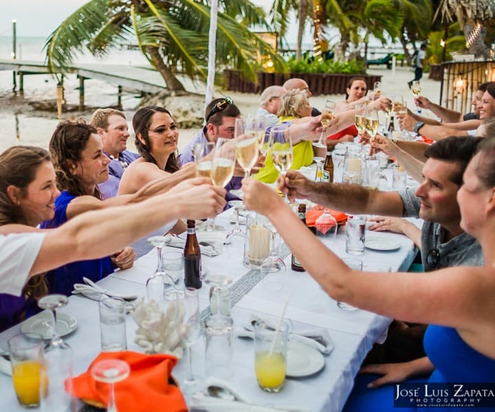 A group of people toasting with drinks at a festive outdoor dinner table by the beach.