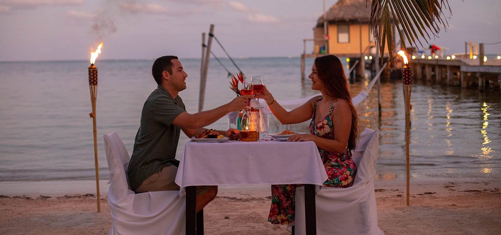 A couple sits at a table on covered in a white tablecloth, toasting with wine glasses. It is twilight. The pier is visible in the background.