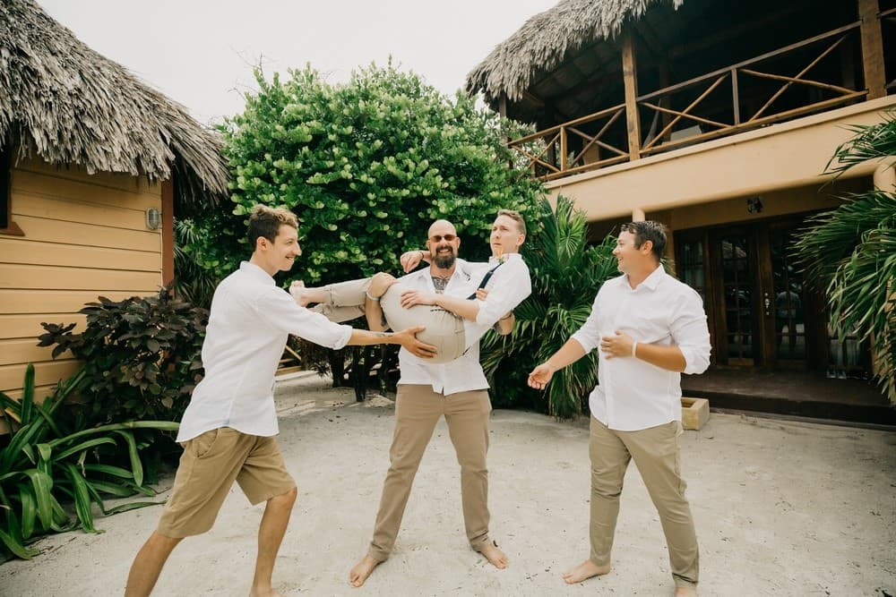 Four men playfully engage outdoors, with one man being lifted off the ground.