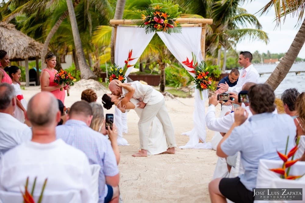 A couple shares a kiss during their beach wedding ceremony surrounded by guests and tropical decor.