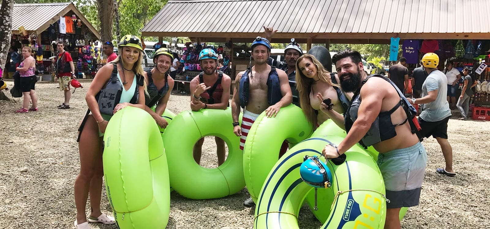 A group of people in swimwear and helmets pose happily with large green inner tubes. The setting is outdoors, near a shop with colorful gear. The mood is joyful and adventurous.
